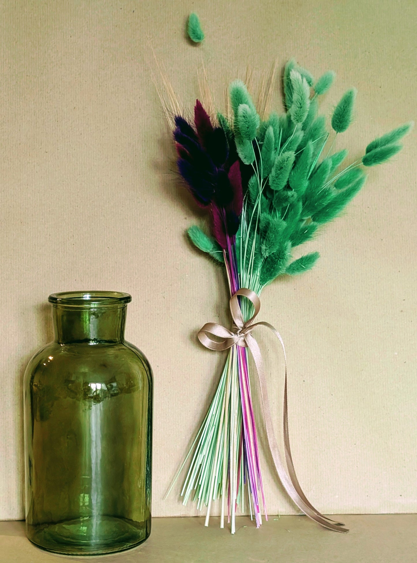 Green vase next to a bouquet of green feathers and ribbons on a beige background