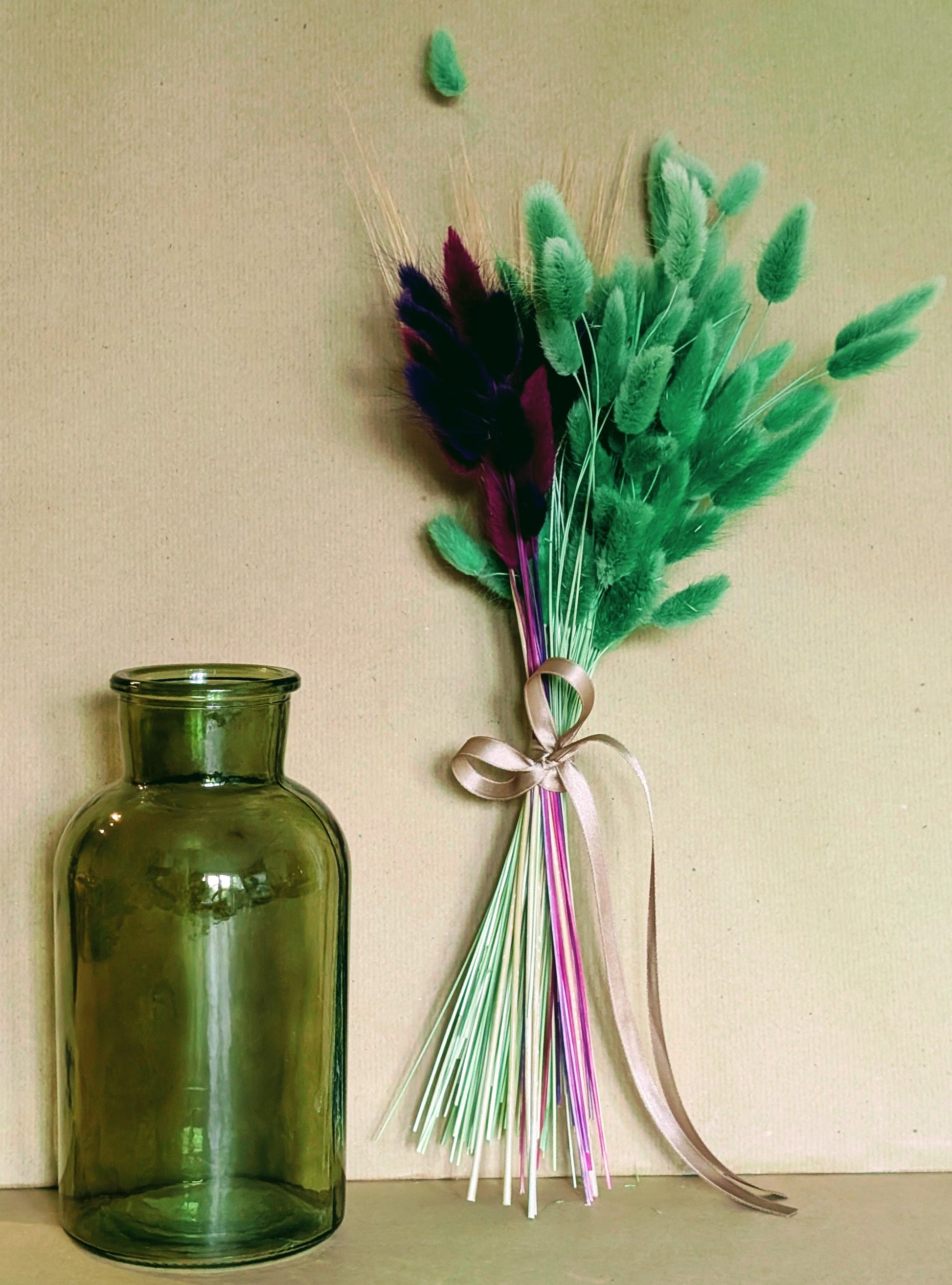 Green vase next to a bouquet of green feathers and ribbons on a beige background